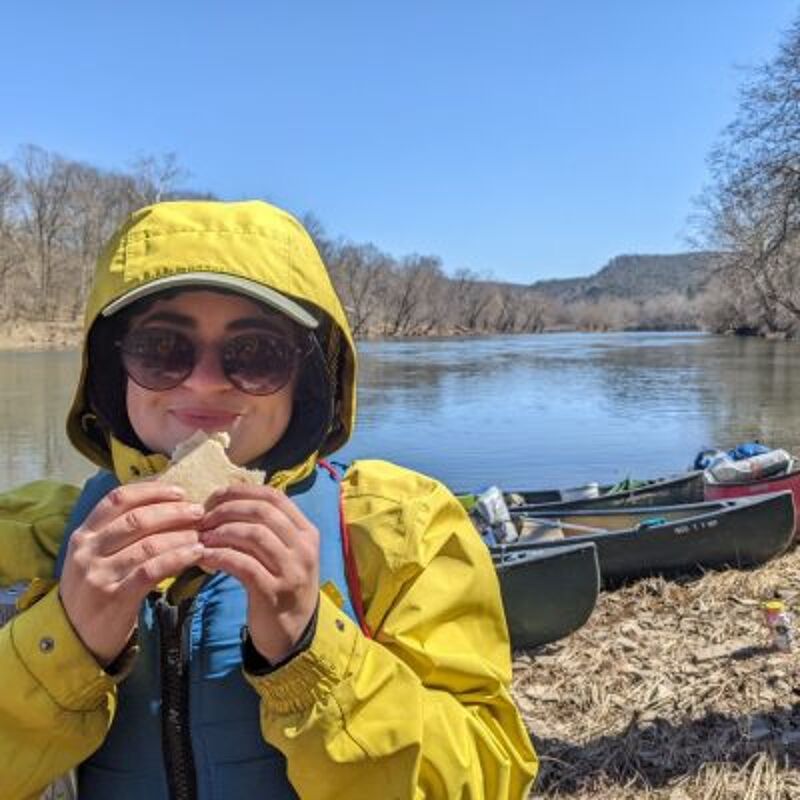 A woman wearing a yellow hooded jacket and sunglasses is eating a sandwich by a river. She is wearing a blue life vest. Behind her, there are two canoes on the riverbank. The river is wide and calm, with trees lining the opposite shore. The sky is blue and clear.